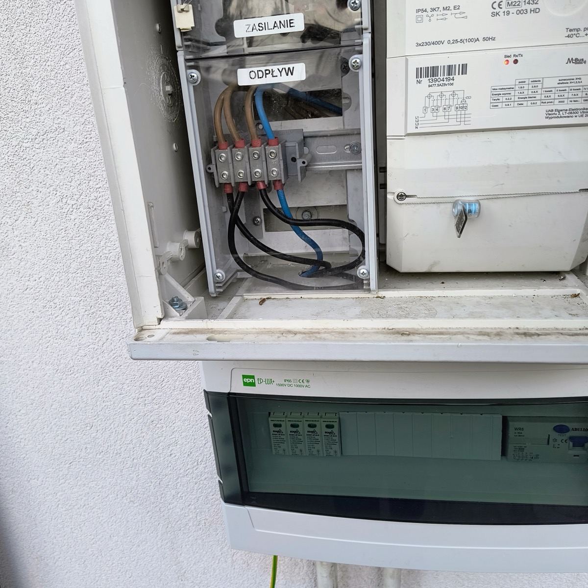 Skrzynka pomiarowo-rozdzielcza na zewnątrz budynku, z licznikiem energii i podłączeniem zasilania