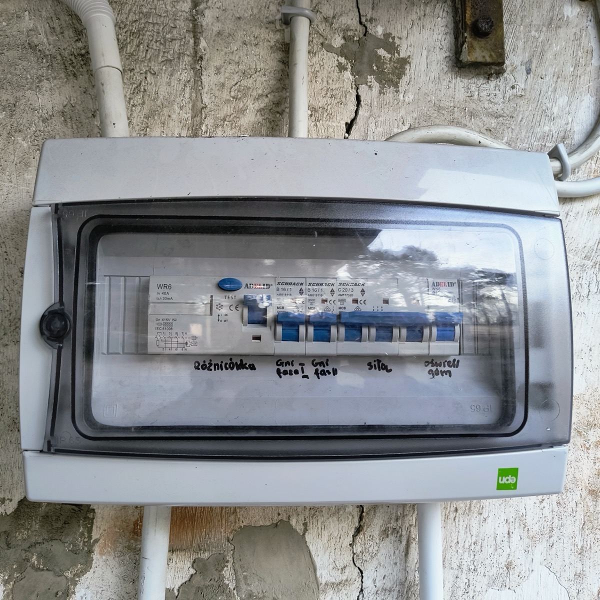 Rozdzielnica elektryczna na zewnątrz budynku z opisanymi obwodami: różnicówka, gniazda, siła, oświetlenie